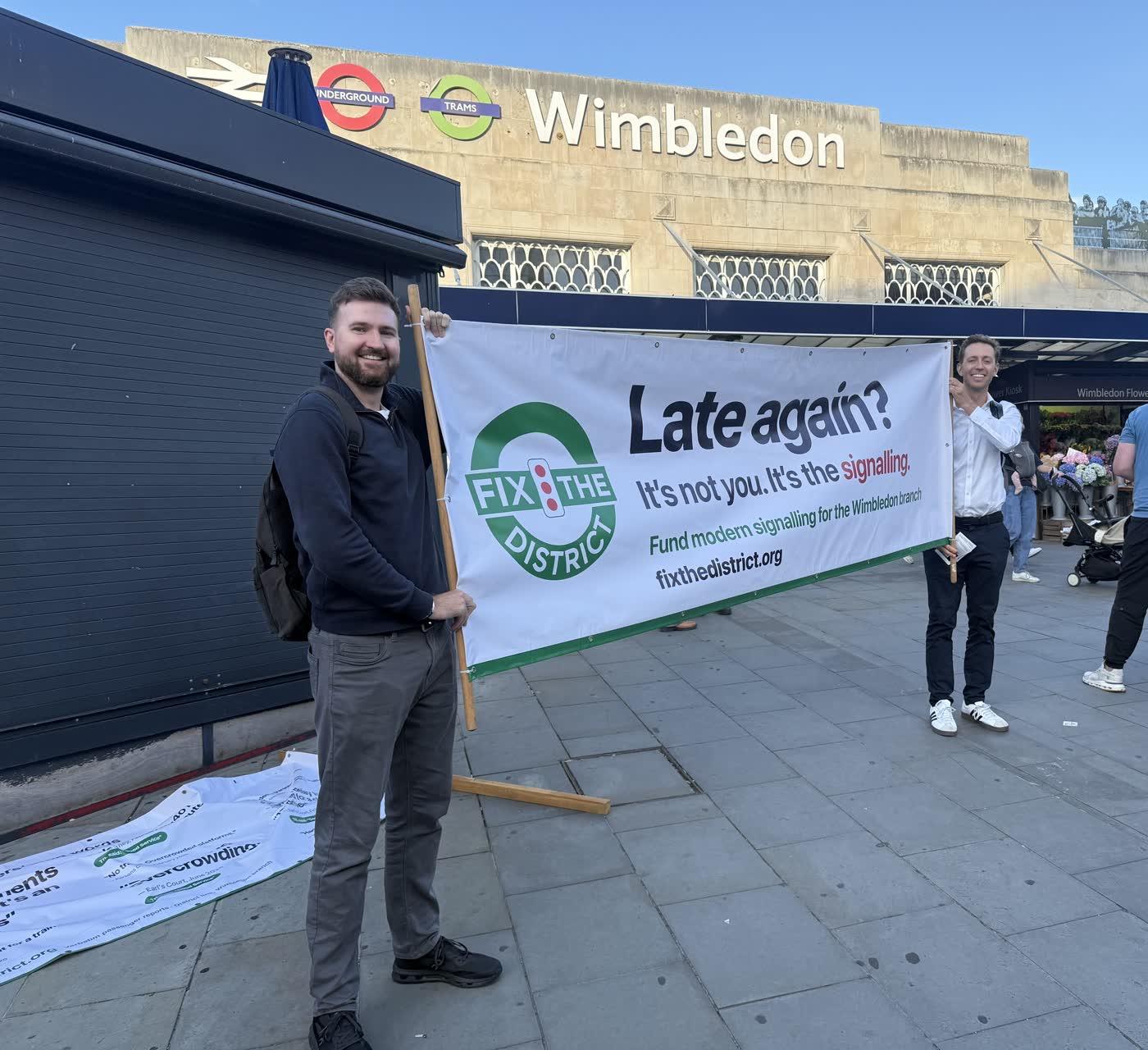 The campaign banner held up along the pavement with the Wimbledon station frontage behind it.