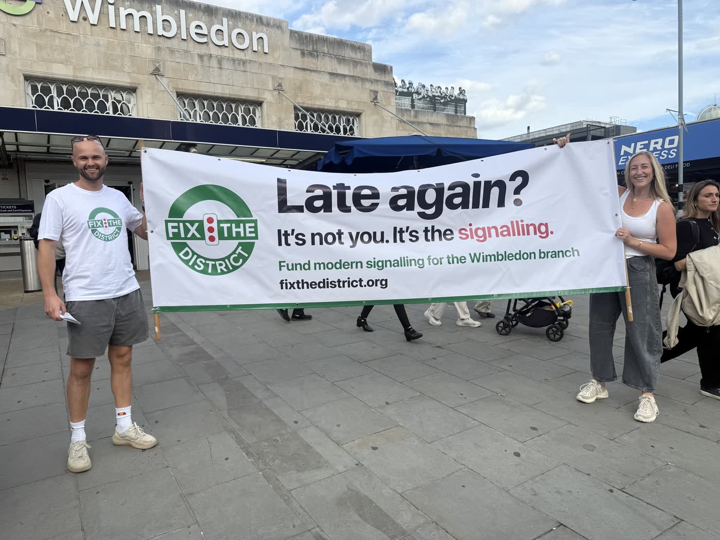 Two campaigners holding the “Late again? It’s not you. It’s the signalling.” banner under the Wimbledon station sign.