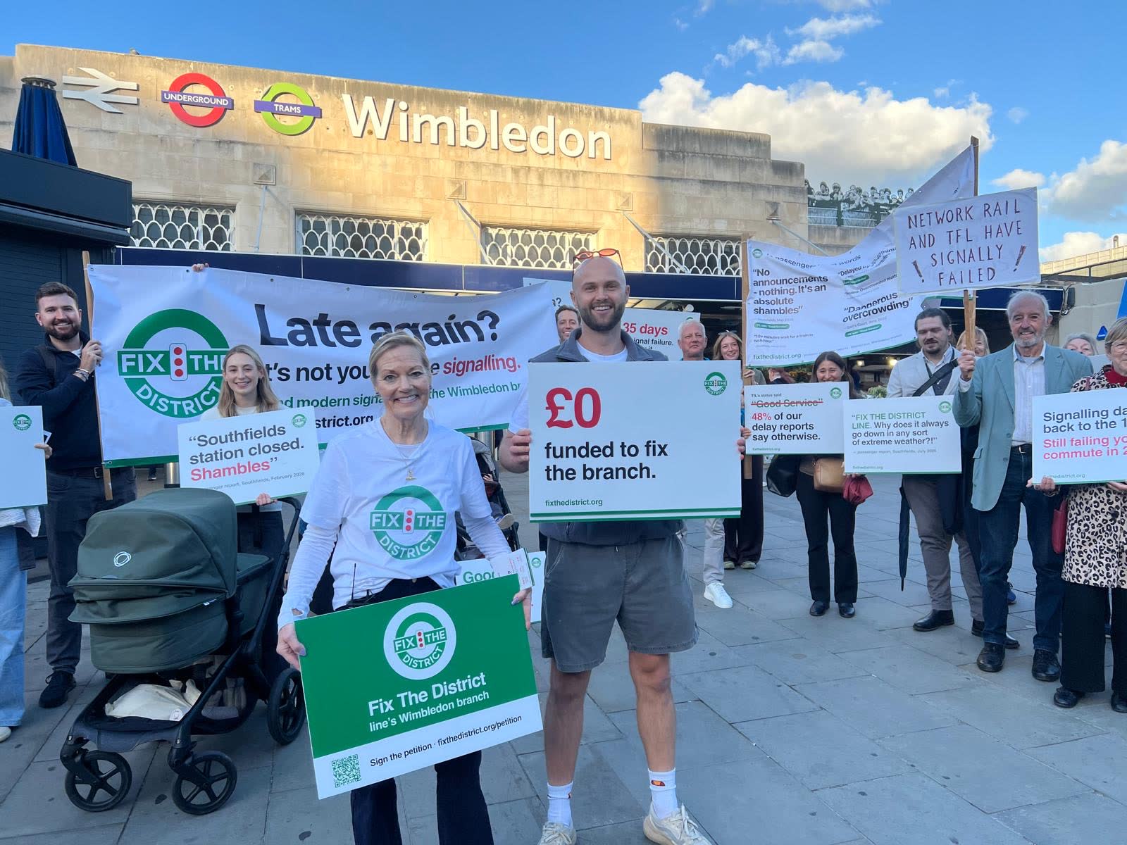 A crowd of passengers outside Wimbledon station holding Fix The District placards and a &ldquo;Late again? It&rsquo;s not you. It&rsquo;s the signalling.&rdquo; banner.