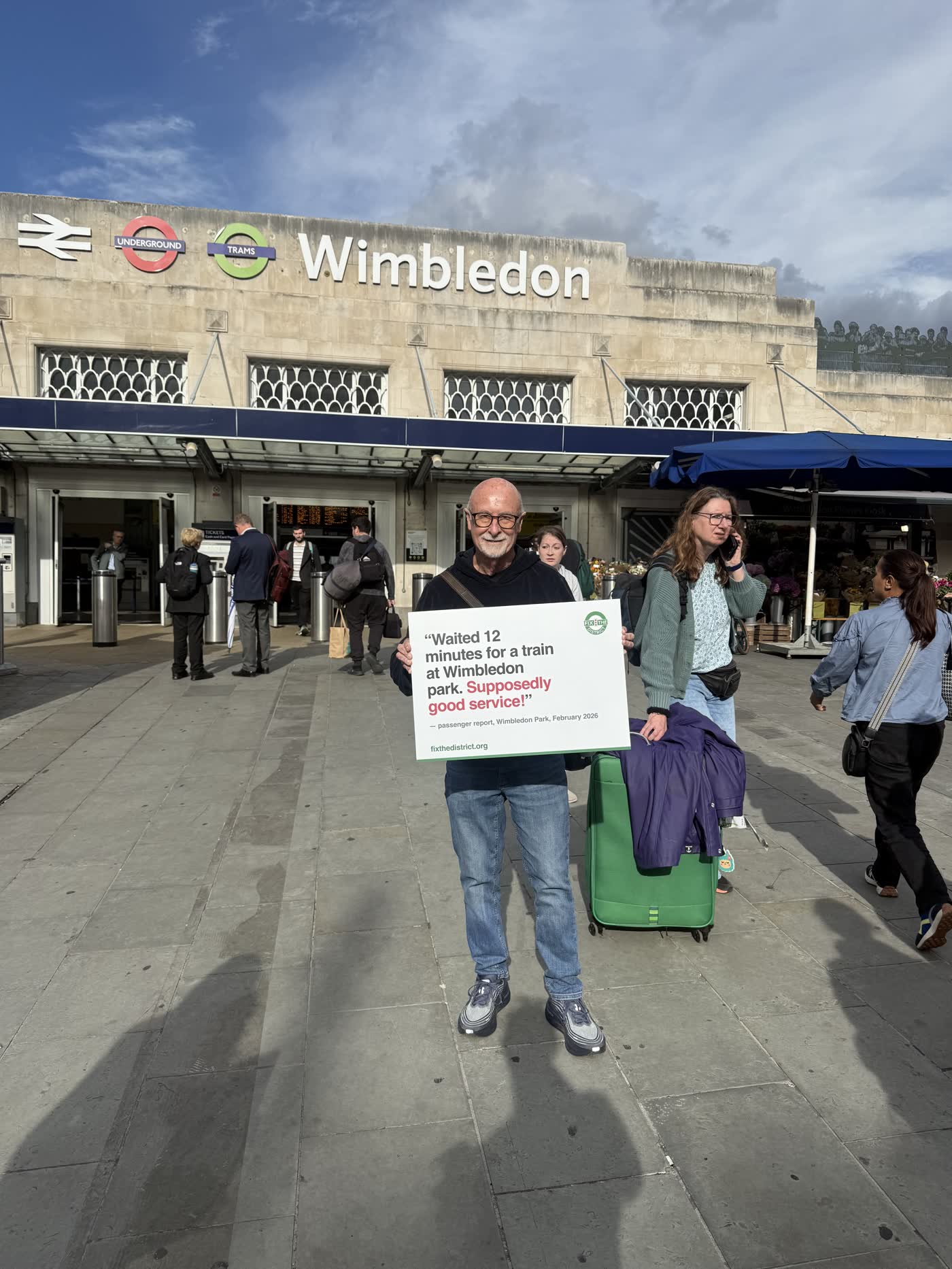 A passenger holding a placard quoting a delay report: “Waited 12 minutes for a train at Wimbledon Park. Supposedly good service!”